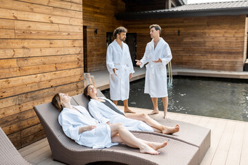 Two female friends lie in bathrobes on sunbeds outdoors after spa treatments and sauna while two...