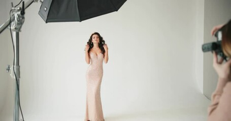 Model posing in the sutdio. Female photographer takes a picture of lady in pink evening dress standing on the white background