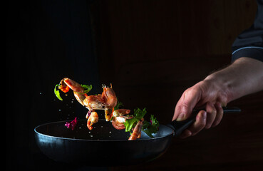 Professional chef cooks shrimp in a pan with vegetables. Cooking seafood, healthy vegetarian food and food on a dark background. Freezing in motion. Free advertising space
