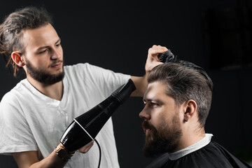 Barber dries the hair of a handsome bearded man after a fashionable haircut. The work of a hairdresser during the quarantine period coronavirus covid-19.