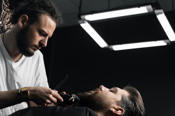 Handsome bearded man has hair and beard cut in barbershop. Dreadlock barber cuts client's hair