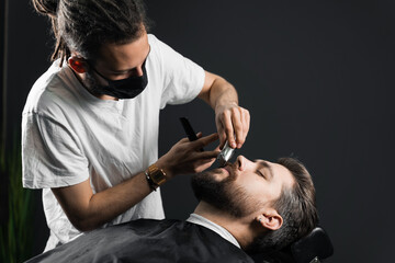 Obraz na płótnie Canvas Mustache shaving in barbershop. Barber with dreadlocks in black medical mask trim beard of handsome man at quarantine coronavirus covid-19.