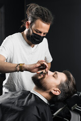 Mustache shaving in barbershop. Barber with dreadlocks in black medical mask trim beard of handsome man at quarantine coronavirus covid-19.
