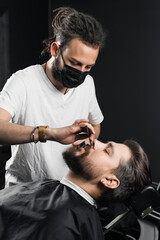 Fototapeta premium Mustache shaving in barbershop. Barber with dreadlocks in black medical mask trim beard of handsome man at quarantine coronavirus covid-19.