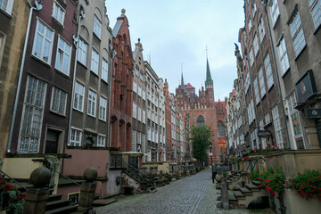 Fototapeta na wymiar A deserted street in Old Town of Gdansk, Poland. The both sides of a paved alley have tall medieval buildings with colorful facades. Bell tower at the end of the alley. A bit of overcast. City tour.