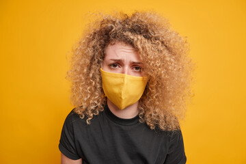 Close up shot of curly haired woman looks sadly at camera wears protective mask being fed up of lockdown restrictions wears black t shirt isolated over yellow background. Coronavirus pandemic