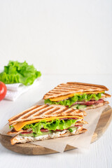 Two homemade organic sandwiches made of cucumber, ham, cheese, lettuce and tomatoes between slices of grilled toasted bread served on cutting board on white wooden table with ingredients. Vertical