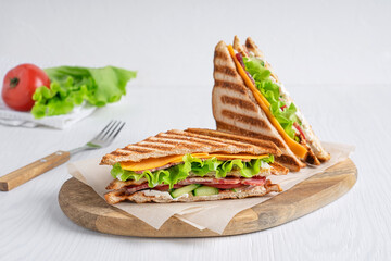 Two homemade sandwiches made of cucumber, slice of meat and cheese, lettuce and tomatoes between slices of grilled toasted bread served on cutting board on white wooden table with knife. Horizontal
