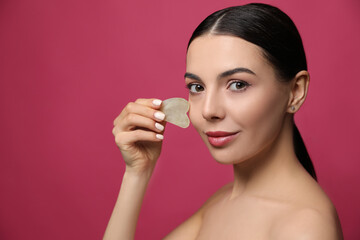 Beautiful young woman doing facial massage with gua sha tool on pink background
