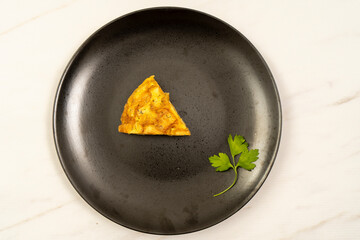 Aerial view of a portion of potato or papa omelette on a black plate on a light marble table. Traditional gourmet tapas concept.