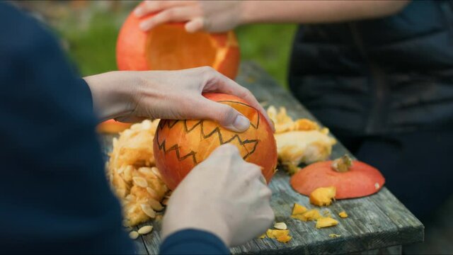 Over the shoulder shot of a caucasian woman carving pumpkins with her pre-teenage son, making jack-o-lantern on the wooden table in the yard
