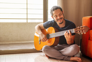 Young latin man sitting on the floor playing acoustic guitar at home in the living room with natural window light