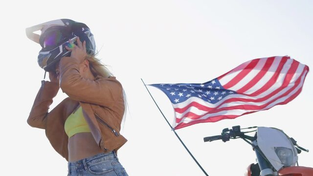 Blonde woman in top and leather jacket puts on helmet by motorbike and waving American flag under clear sky low angle shot