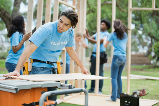 Young man cutting wood with table saw and volunteers building frame in background