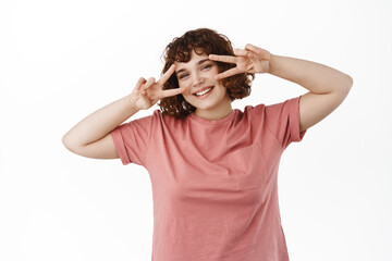 Happy attractive young woman showing v-sign peace kawaii gesture near eyes, dancing disco, smiling and express positivity with joy, standing over white background