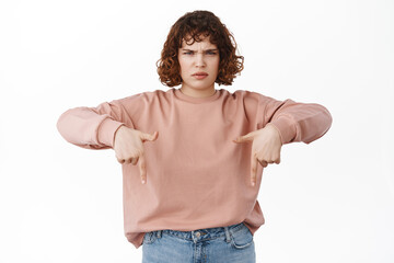 Angry frowned young girl complains, pointing fingers down with sulking upset face, disappointed by...