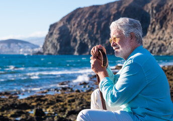 Bearded senior man sitting at sunset light on the beach holding a walking stick because of body aches, looking at horizon over water