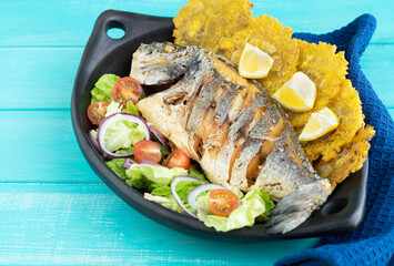 Fried fish with salad and patacones on black plate on blue wooden background. Copy space.