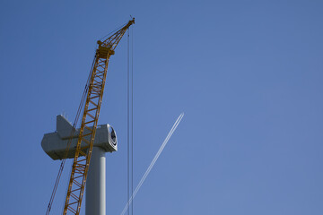 An passenger aircraft is drawing contrails behind a wind turbine under construction