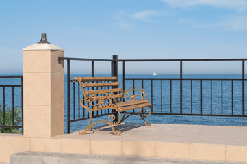 A bench made of wrought metal and wood on the seaside promenade overlooking the sea. A white yacht can be seen in the distance.