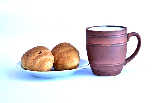 The picture shows a glass made of brown clay with milk poured into it, and next to it there are two buns on a plate.