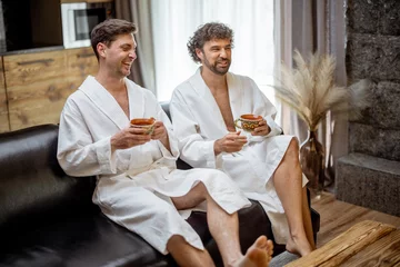Abwaschbare Fototapete Spa Two man in bathrobe sitting on sofa in living room of spa house and drink a tea. Relax and wellness concept. Spending and enjoy time together.