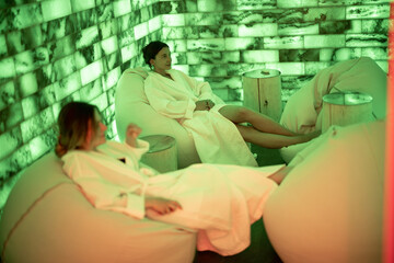 Two women in bathrobe sitting and relaxing in salt room with green light. Health and wellness spa. Leisure time together.