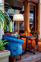 outdoor cafe terrace with soft colored armchairs and round tables on wooden flooring by the window with an entrance door and flowers with a decorative floor lamp, nobody.