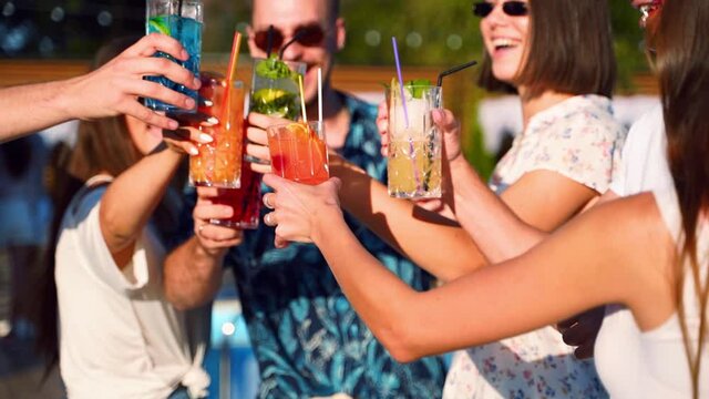 Close view of friends having fun at summer pool party, clinking glasses with cocktails and dancing near hotel swimming pool outdoors. People toast drinking juice at luxury villa in slow motion.