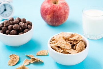 Bowls with cereal and chocolate balls, a glass of milk, an apple and an alarm clock on a blue background. Scheduled breakfast. Close-up