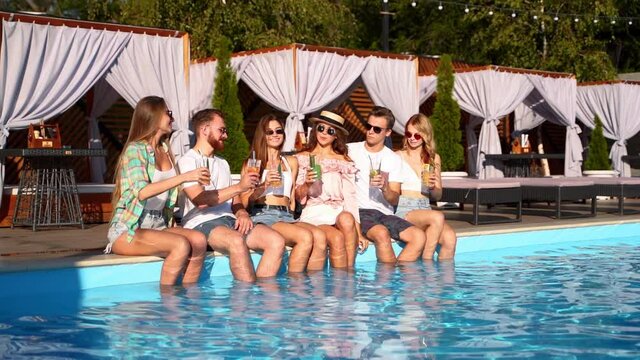 Group of friends having fun at poolside party clinking glasses with fresh cocktails sitting by swimming pool on sunny summer day. People toast drinking beverages at luxury villa on tropical vacation.
