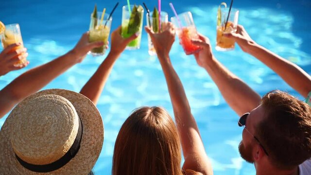 Back view of friends clinking glasses with cocktails sitting by swimming pool on sunny summer day. People have fun at poolside party toasting, drinking beverages at luxury tropical villa. Slow motion.