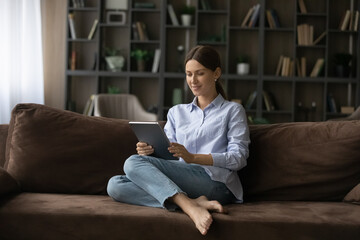 Relaxing with pad. Happy young lady spend time on comfy sofa at home hold digital tablet chat...