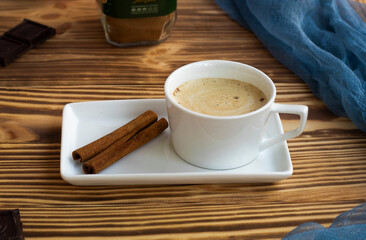 a cup of latte with froth stands on a square saucer on a wooden table. Cinnamon sticks, dark...