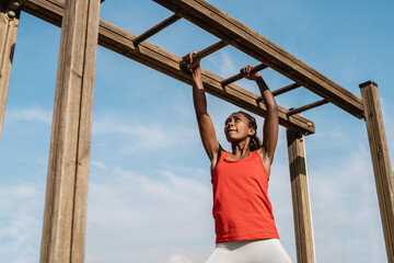 African young woman climbing monkey bars in military training boot camp outdoors at city park -...
