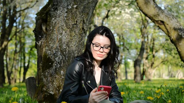 Young woman sitting on a green lawn in the summer park with a smartphone