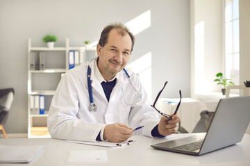 Fototapeta na wymiar Friendly senior male doctor sits in his office in front of a laptop and fills out medical records. Portrait of a doctor sitting at his workplace and looking at the camera. Concept of medical work.