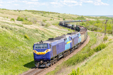 A combination of two diesel locomotives pulls a long train of wagons loaded with products along the winding railways of the spring steppe.  Sunny weather.  Zoom photo
