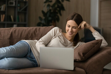 Fototapeta premium Interested millennial lady rest on couch with laptop read blog write comment to social network chat with mate online by wireless internet. Happy young female lying down on sofa working from home on pc