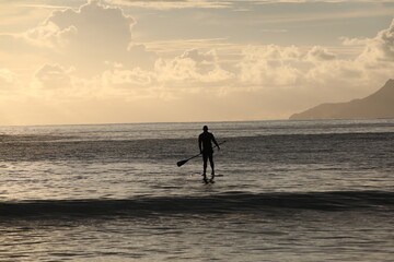 The silhouette of a man going out to sea on a paddle board at dawn.Calm landscape of water with a small wave and an island on the horizon in a light haze.The concept of sports and outdoor activities