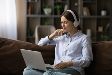 Comfort in learning. Calm young woman sit on cozy couch wear headphones hold laptop watch educational video internet channel study virtual lesson. Smiling teen female in earphones enjoy movie online