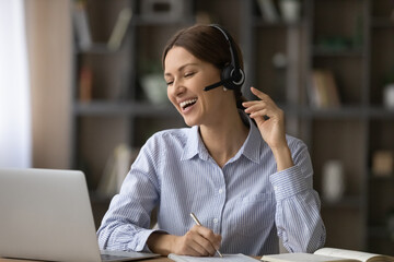 Funny remote learning. Joyful female student teenager taking notes of online virtual lesson laugh on good teacher speaker joke. Young woman in headset microphone having fun handwriting distant lecture - obrazy, fototapety, plakaty