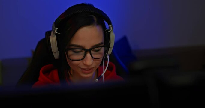 Portrait, cute girl blogger looks at the monitor screen and smiles, she is broadcasting online. Beautiful girl in headphones with a microphone sits in a dark room at a computer, she gestures and