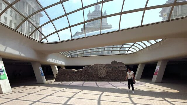 Woman tourist underground Serdica, glass dome and ancient historical ruins of the roman city, with view of the National Assembly building in the background. Slow motion