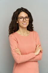Fototapeta premium Portrait of smiling casual businesswoman standing with folded hands over gray wall. Confident business lady or teacher in eyeglasses and pink pullover. Millennial lady positive with curly black hair