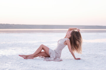 Fototapeta na wymiar Young blonde woman in an evening airy pastel pink powdery dress sitting barefoot on white crystallized salt. Girl with natural make-up, hair is developing. Salt mining trip, walking on water at sunset