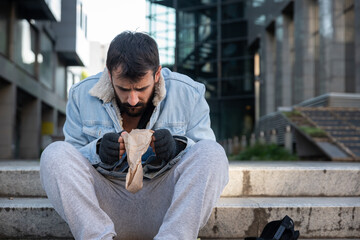 Obraz premium Young hungry dirty depressed homeless man sitting on the stairs of the building on sidewalk and try to eat food scraps from paper bag that he find in the garbage can, social documentary concept