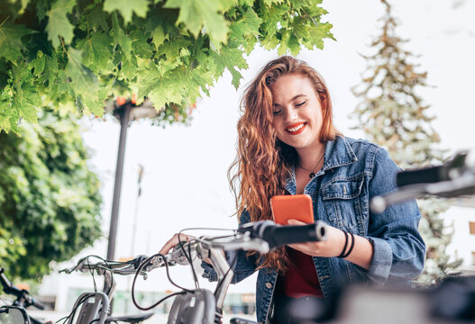 Portrait of smiling red curled long hair caucasian teen girl block out bike at Bicycle sharing point using the modern smartphone. Green urban transport and modern technology devices concept image.