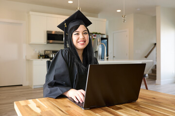 Smiling Asian woman in graduation gown sitting in kitchen at table with laptop, attending virtual...