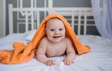 smiling baby boy in a diaper with an orange towel on his head lies on the bed in the bedroom and...